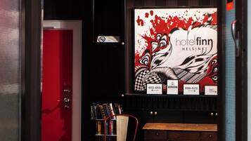 Entrada de hotel con un letrero de neón que dice “sweet dreams”, una recepción, una puerta roja y una pared con un gráfico e información de contacto.