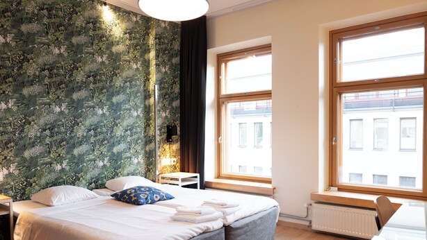 Ett modernt hotellrum med en stor säng, två sängbord, ett litet bord och utsikt över staden genom stora fönster.