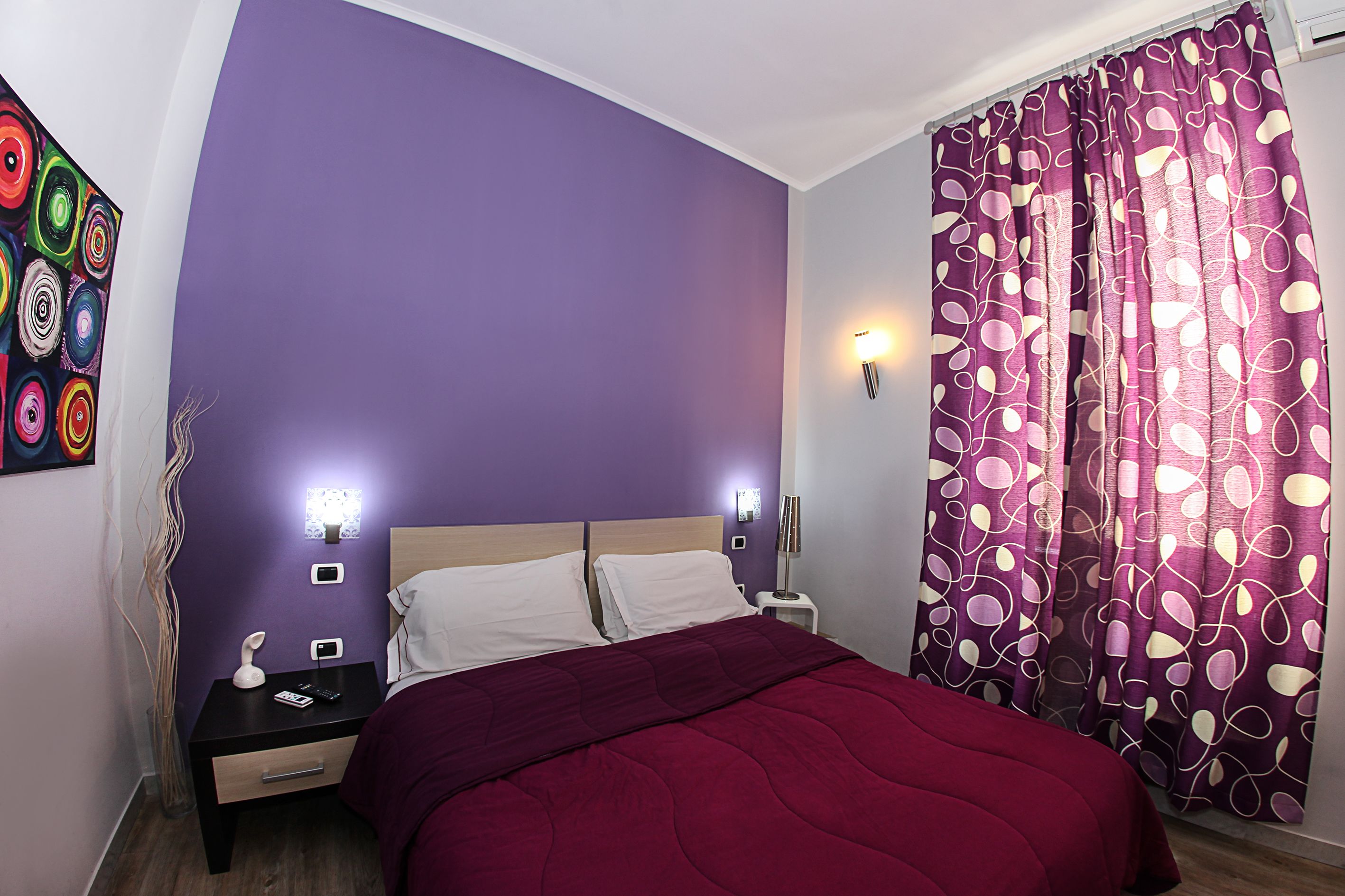 Un dormitorio con pared morada, una cama con manta roja, dos mesitas de noche con lámparas y una cortina con estampado de hojas.