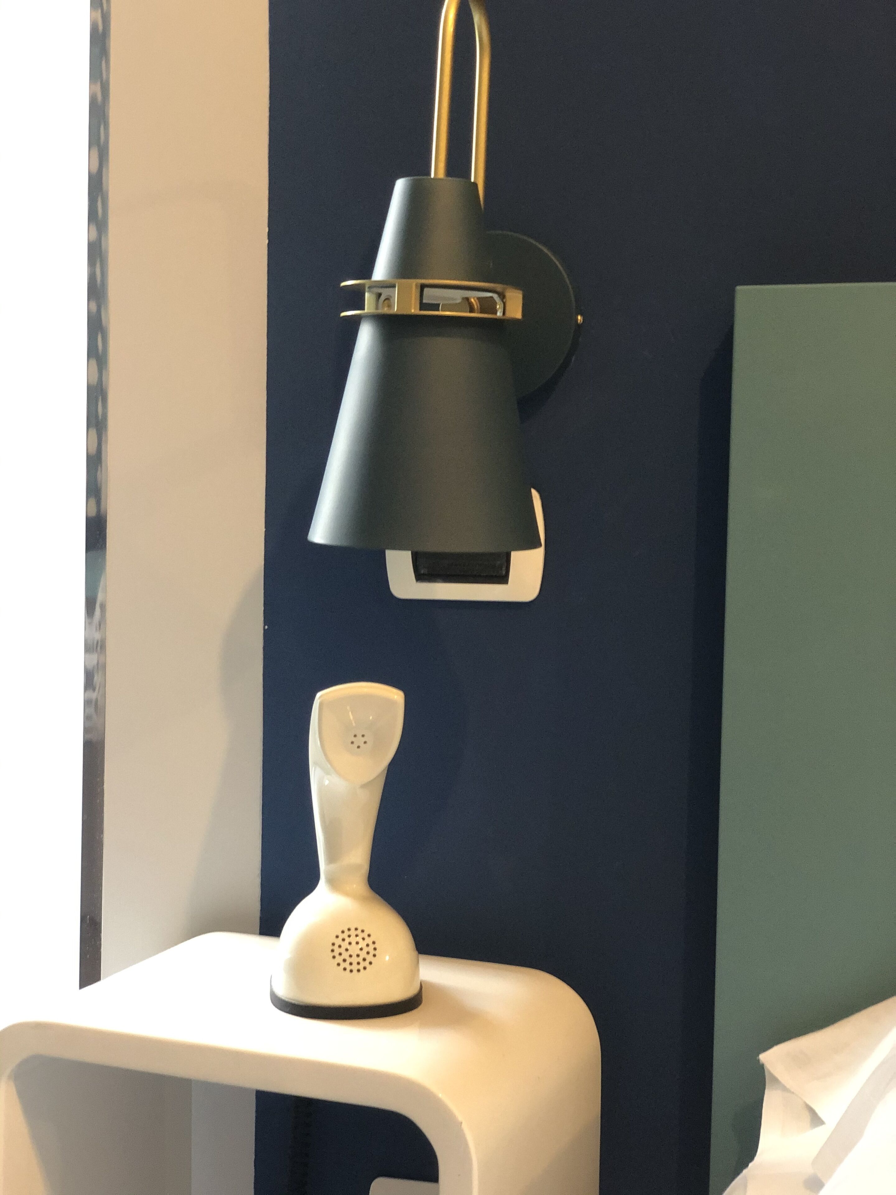 Una lámpara de escritorio blanca con base blanca y pantalla negra, ubicada sobre un escritorio blanco frente a una pared azul oscuro.
