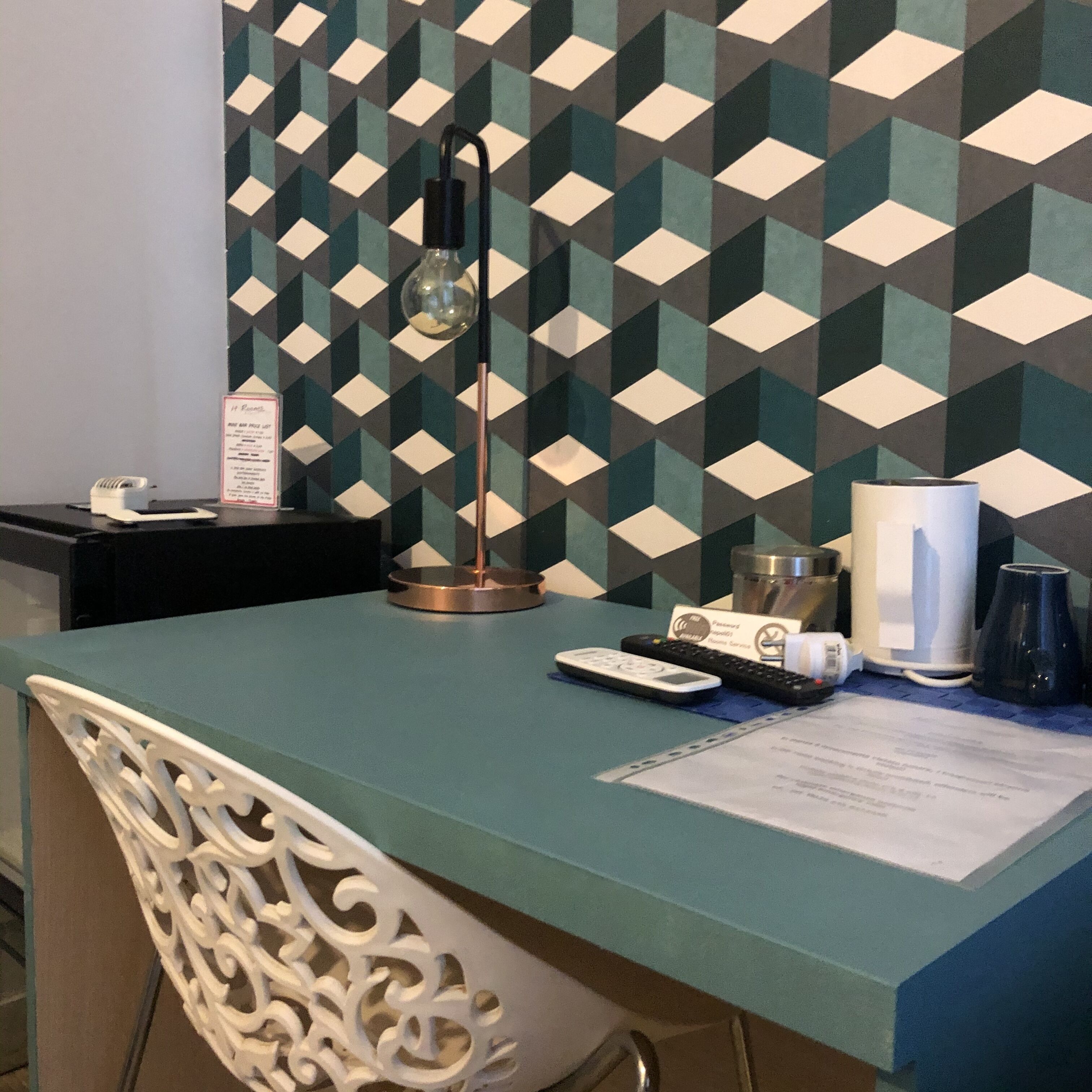A hotel room with a geometric patterned wall, a desk with a lamp, a coffee maker, a remote control, and a decorative chair.