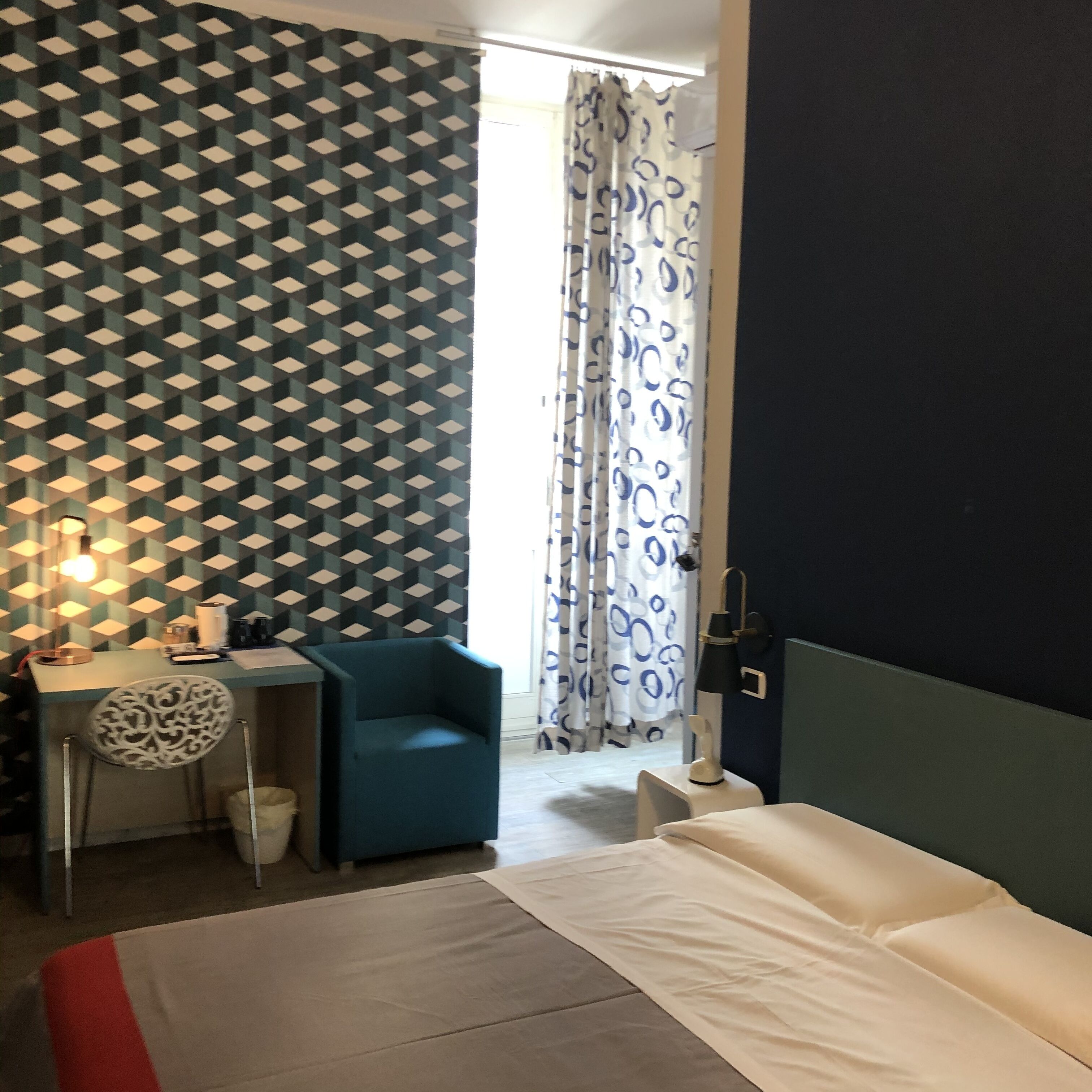 Quarto de hotel com cama, escrivaninha, cadeira e parede estampada.
