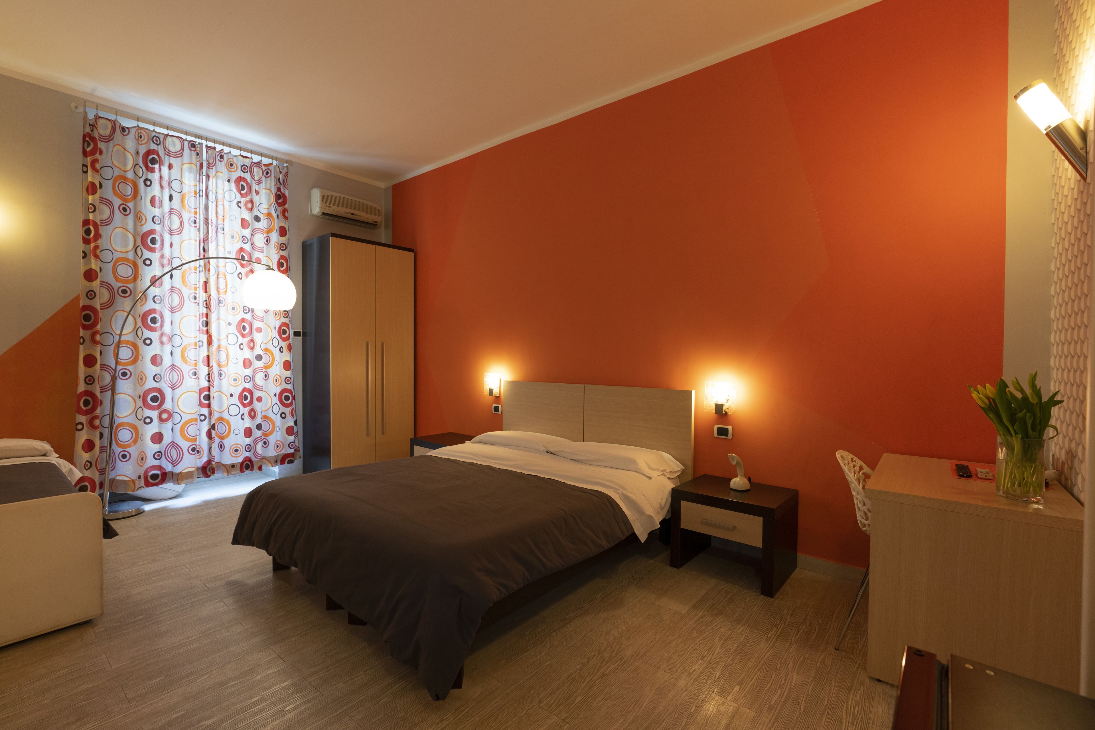 A hotel room with a bed, bedside tables, a desk, a chair, and a curtain with a circular pattern.