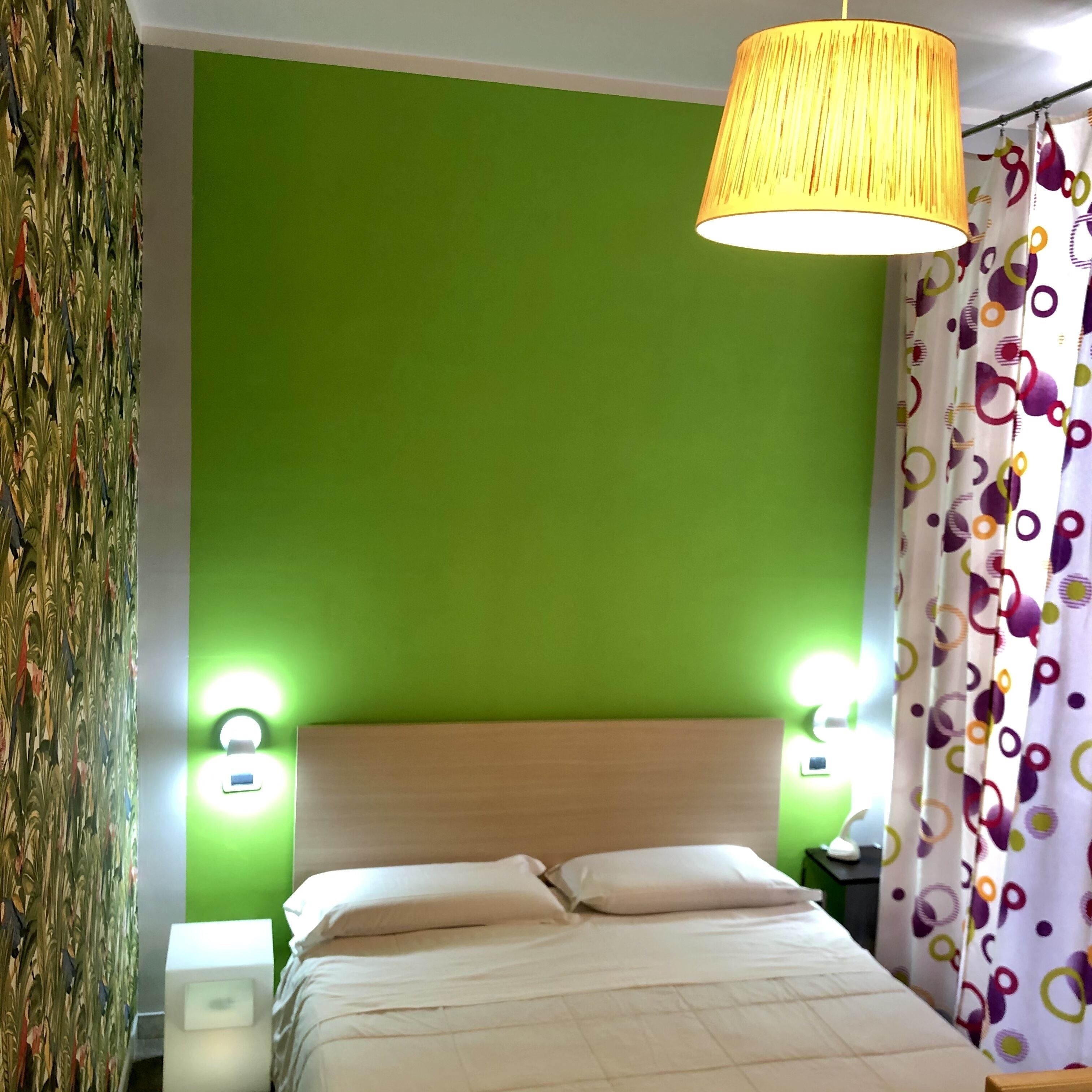 Una habitación con pared verde, cama con ropa blanca, lámpara amarilla y cortina estampada.