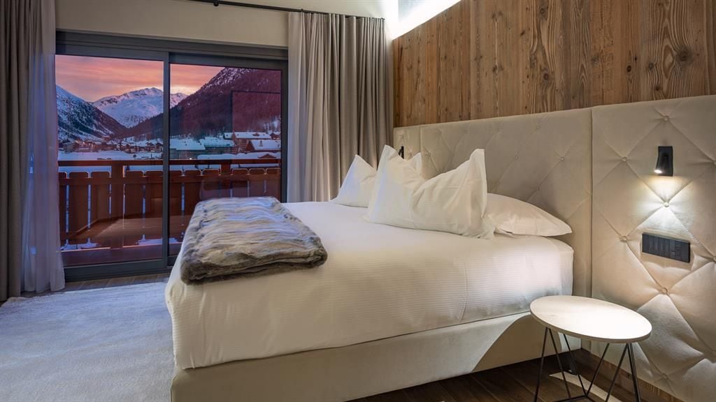 A hotel room with a large bed, a small table, and a view of a snowy mountain landscape through a sliding door.