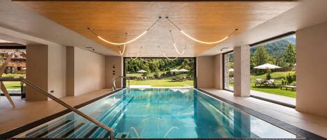 Una piscina interior moderna con paredes de vidrio transparente, techo de madera y vistas a un jardín ajardinado.
