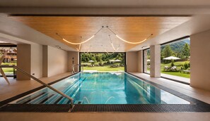 A modern indoor swimming pool with clear glass walls, a wooden ceiling, and a view of a landscaped garden. - Paradise Lodge (Livigno)