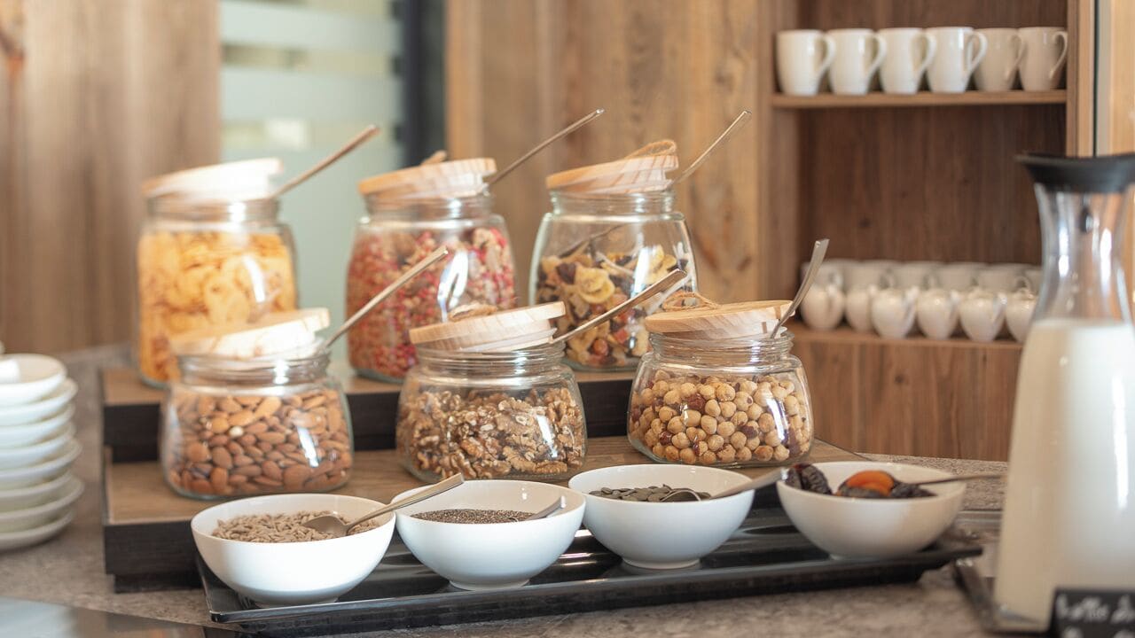 Un bufé de desayuno con diferentes alimentos en frascos y recipientes, una cafetera y una bandeja de platos.