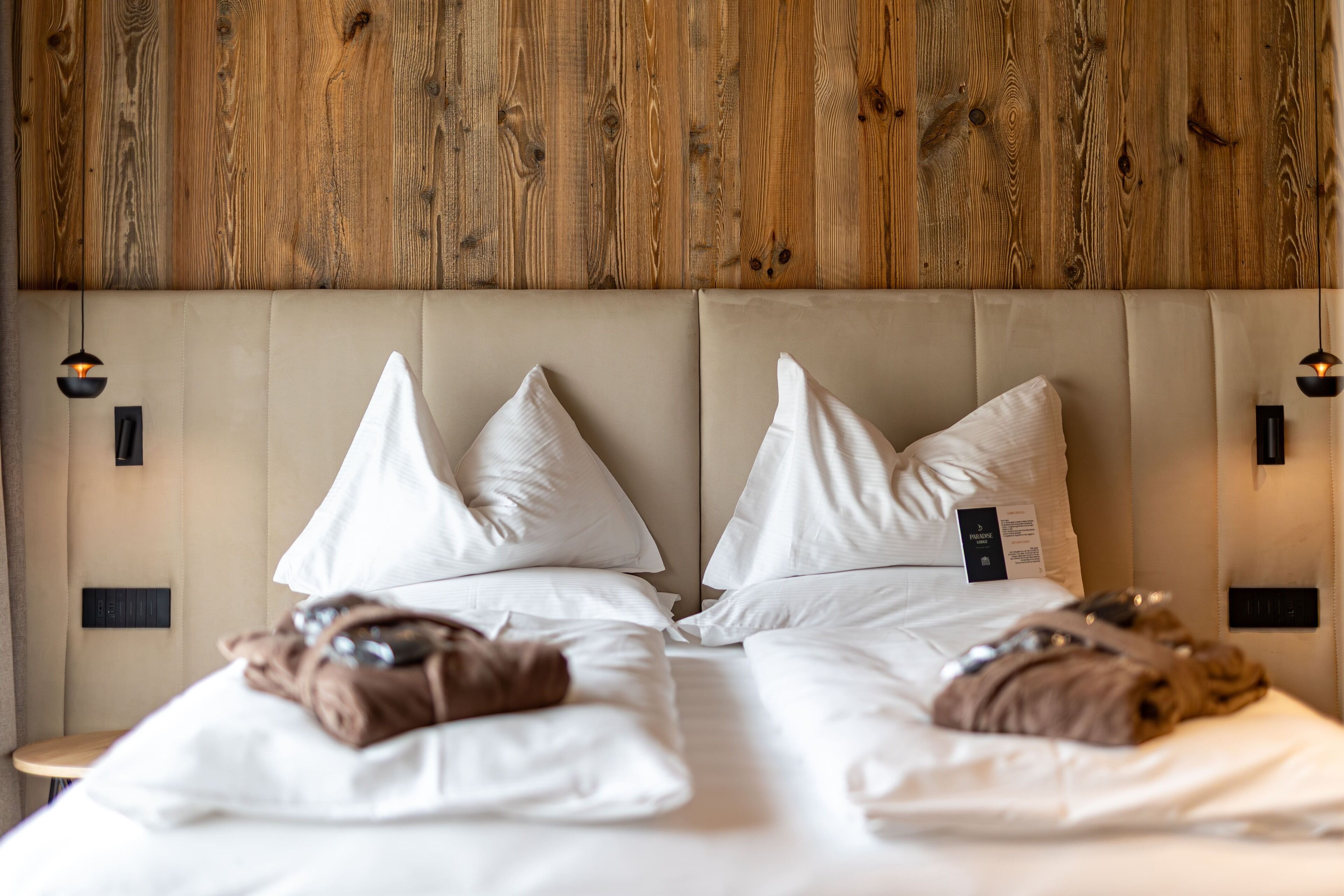 Quarto de hotel com cabeceira de madeira, duas camas com roupa de cama branca e duas luminárias fixadas na parede.