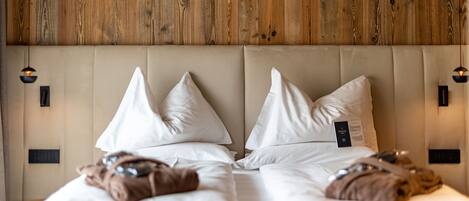 Quarto de hotel com cabeceira de madeira, duas camas com roupa de cama branca e duas luminárias fixadas na parede.