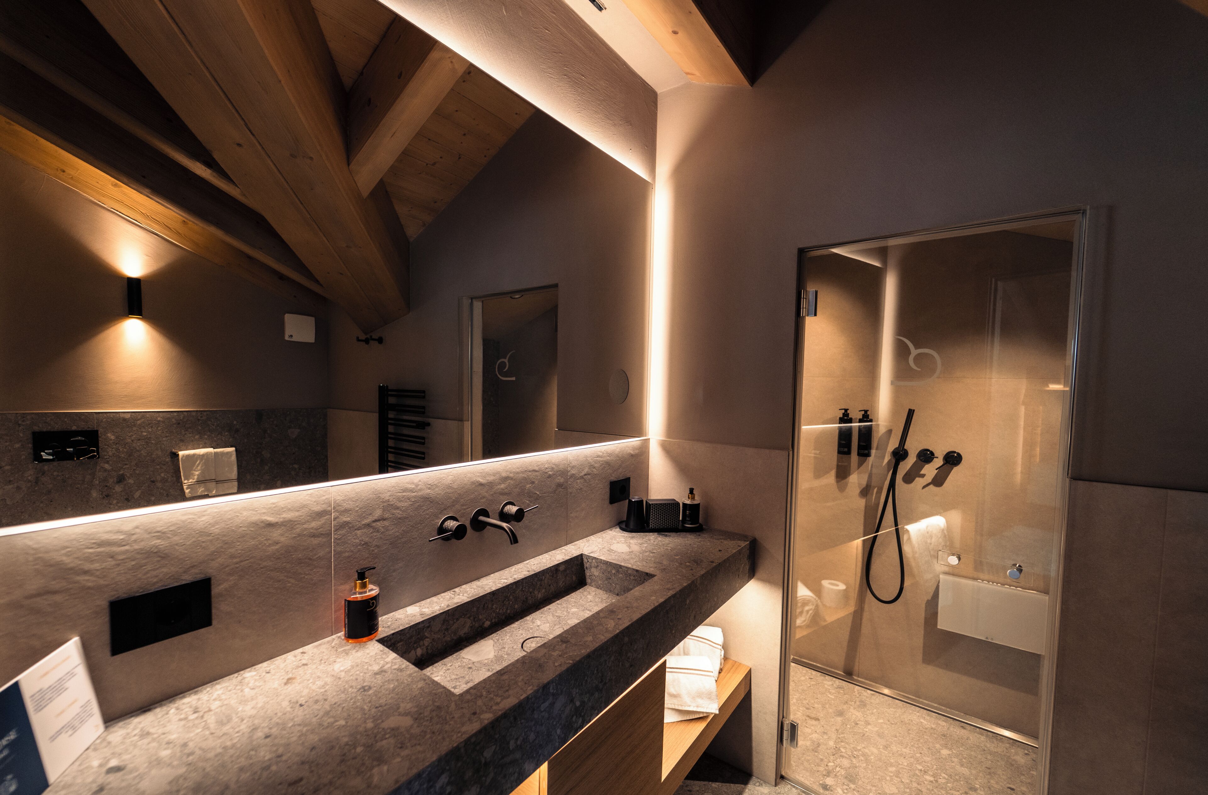 Un baño moderno con un espejo grande, encimera de granito, cabina de ducha y vigas de madera en el techo.
