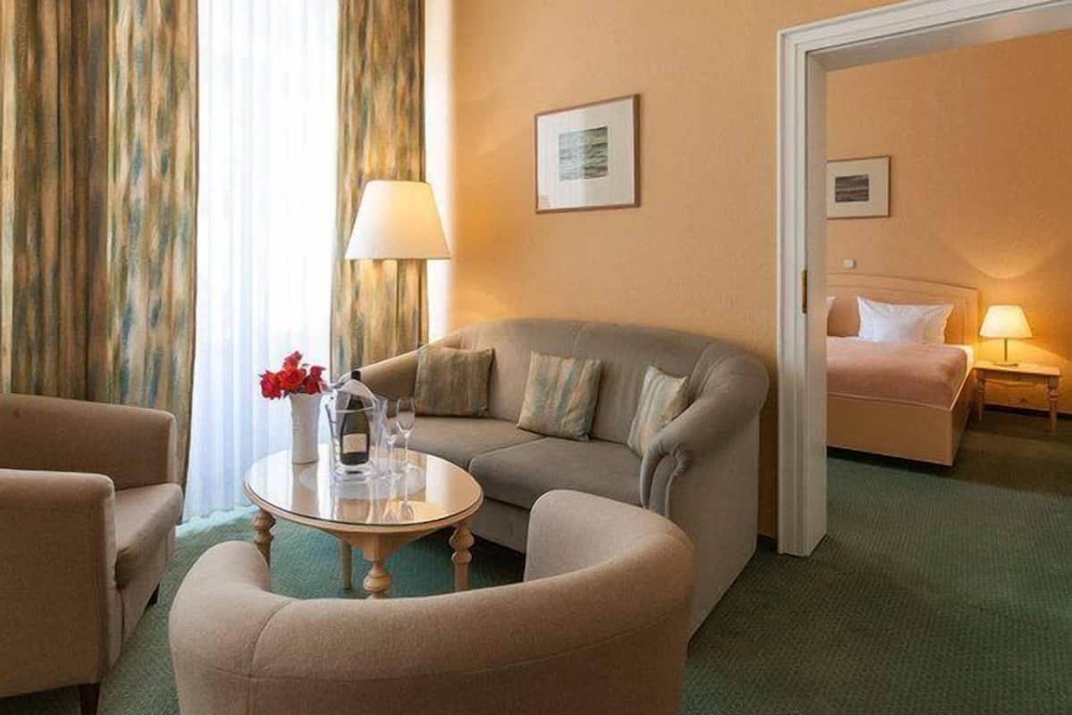 Ein Hotelzimmer mit Sofa, Sessel und einem kleinen Tisch mit Blumenvase. Es gibt ein Bett mit Nachttisch und Lampe.