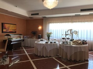A dining area with round tables covered in white tablecloths, a buffet station, and a large window with sheer curtains.