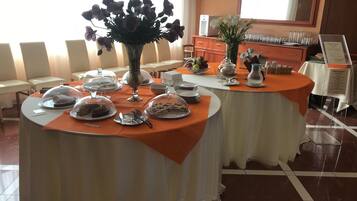 Ein Essbereich mit runden Tischen, bedeckt mit weißen Tischdecken und orangefarbenen Tischdekorationen, zeigt eine Auswahl an Desserts und eine Blumenvase.