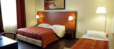 A hotel room with a large bed, a smaller bed, a desk, and a chair. There are two bedside lamps and a painting on the wall.