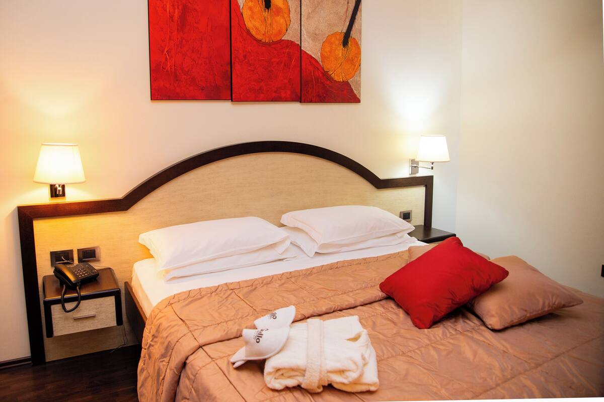 A neatly made bed with white and red pillows, a folded towel, and a robe on the bed. There is a nightstand with a lamp and a phone on it. The room has a wooden headboard and a red abstract painting on the wall.