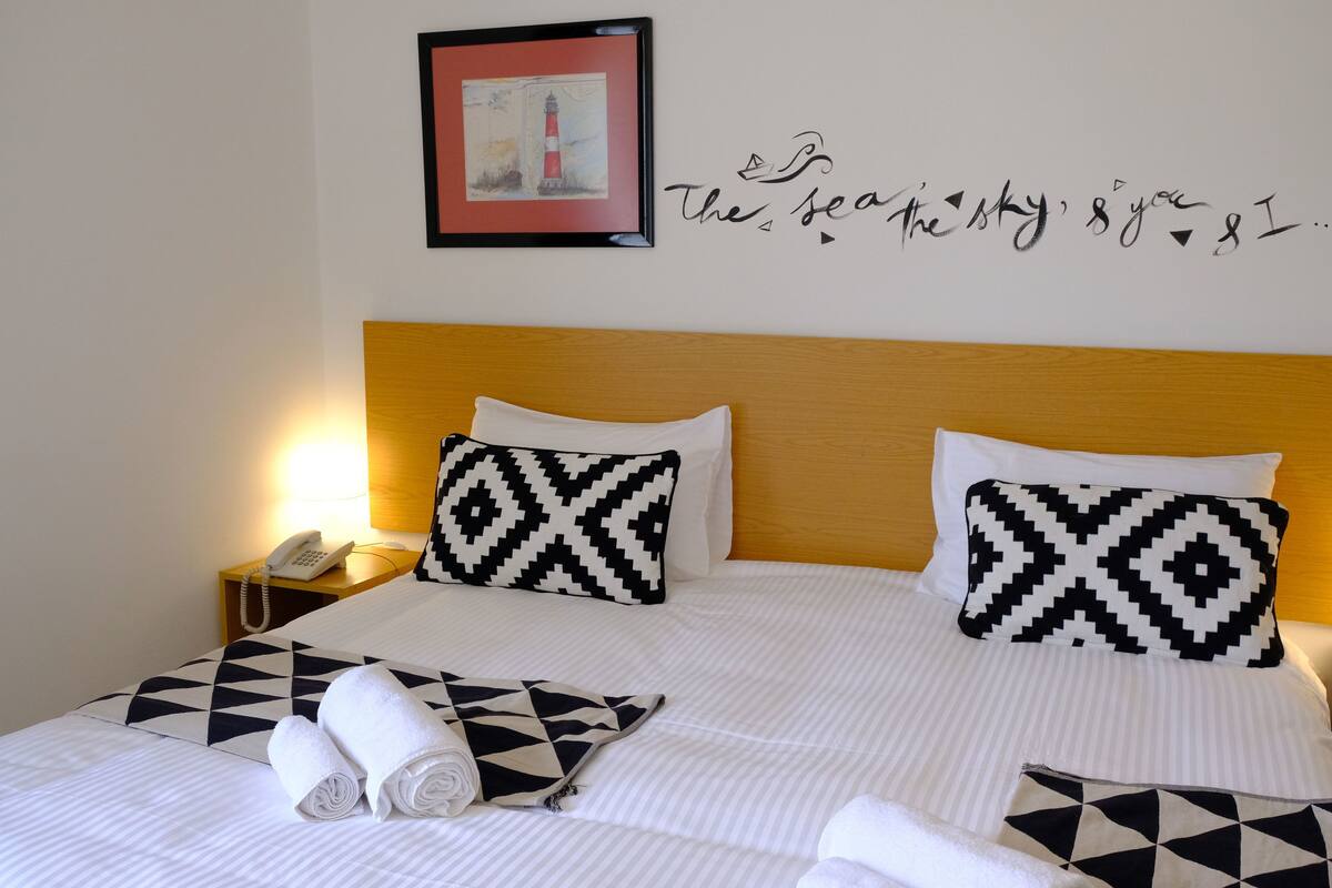 Ein ordentlich bezogenes Bett mit gemusterten Kissen, ein gerahmtes Kunstwerk an der Wand und eine Wandsticker mit einer Botschaft.