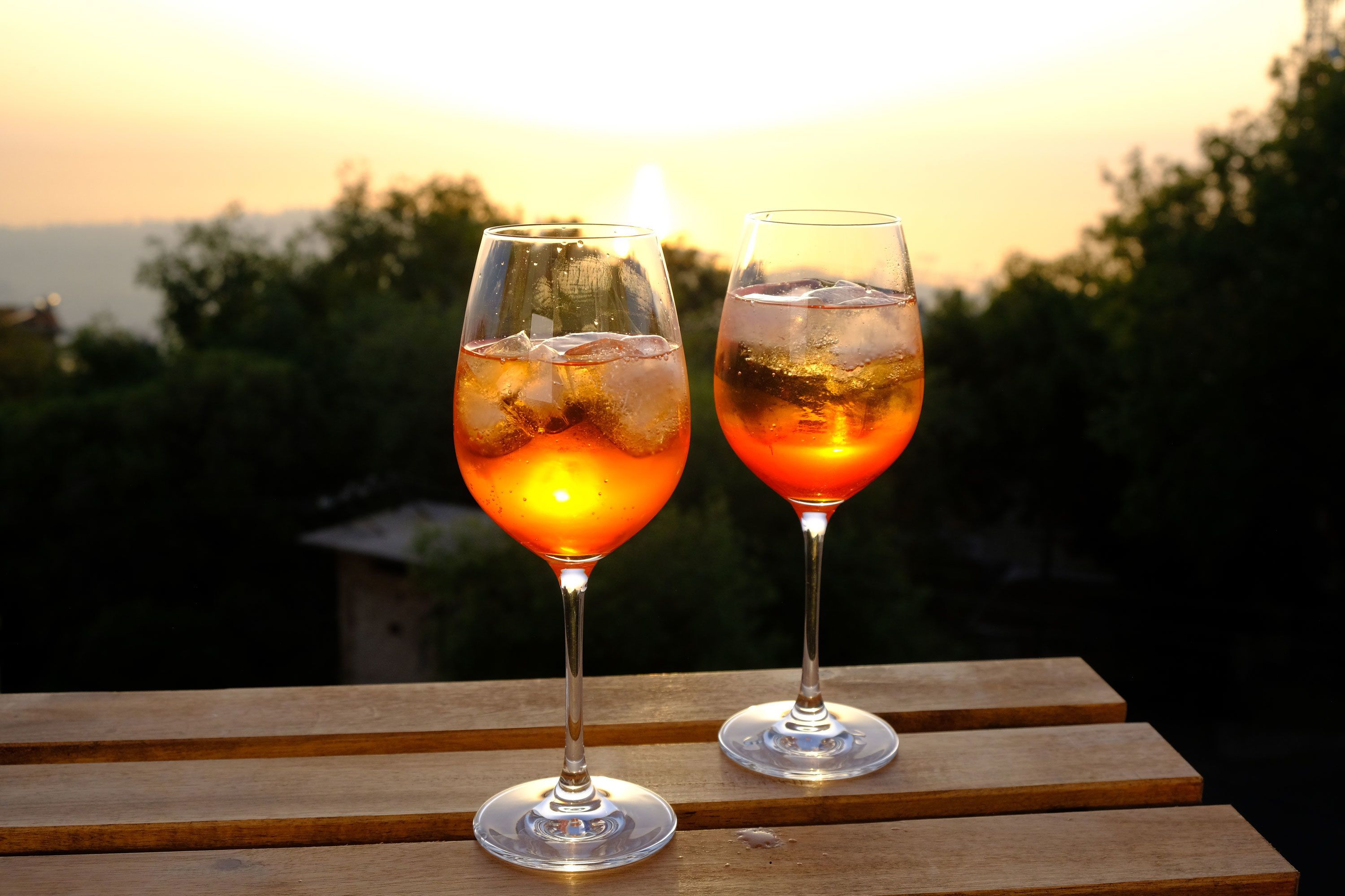 Twee glazen van een drankje op een houten ondergrond, met een zonsondergang op de achtergrond.