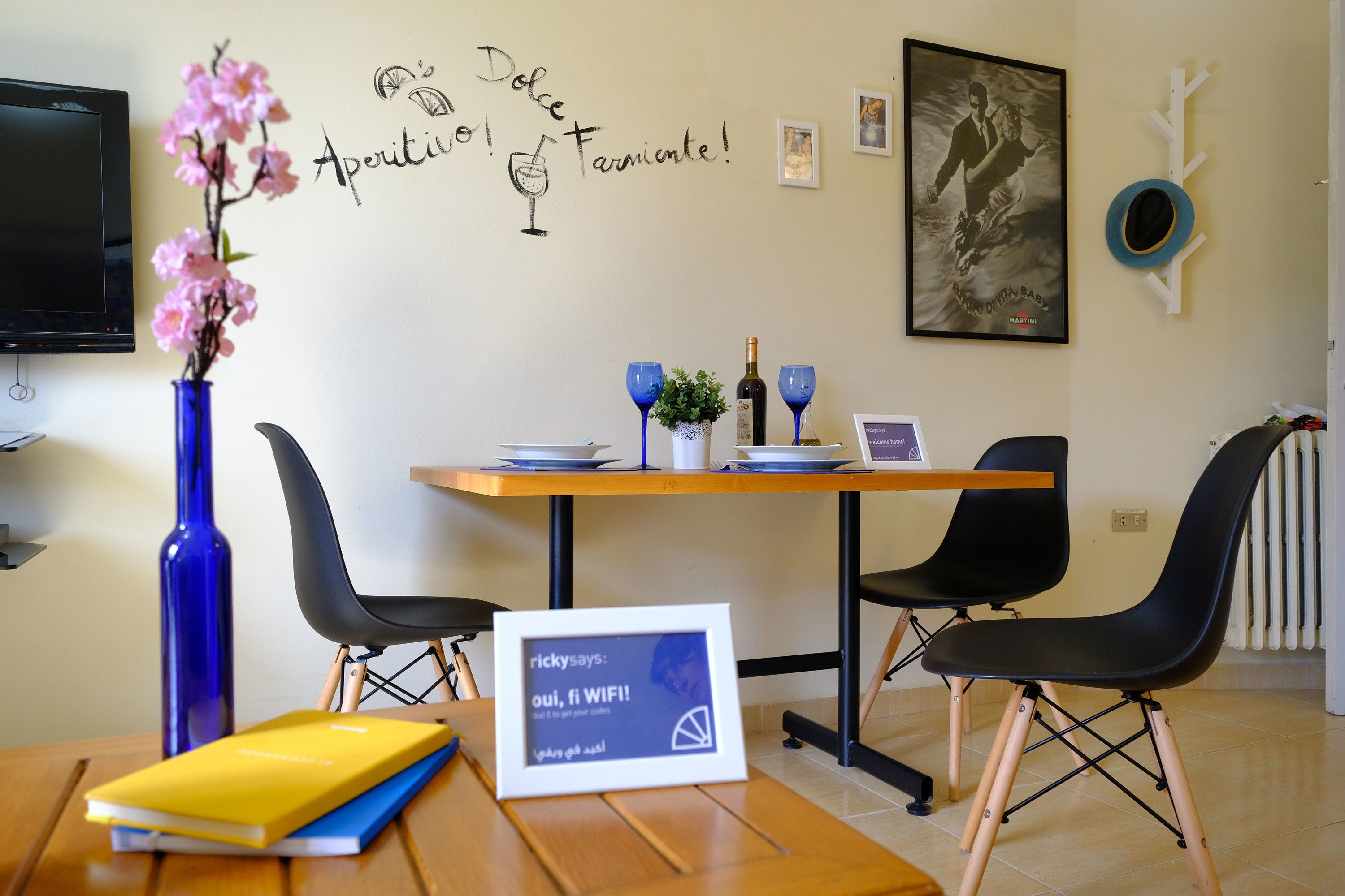 Een eetgedeelte met een tafel, stoelen, een vaas met bloemen, een lijst met een foto en een bord met een computerscherm.