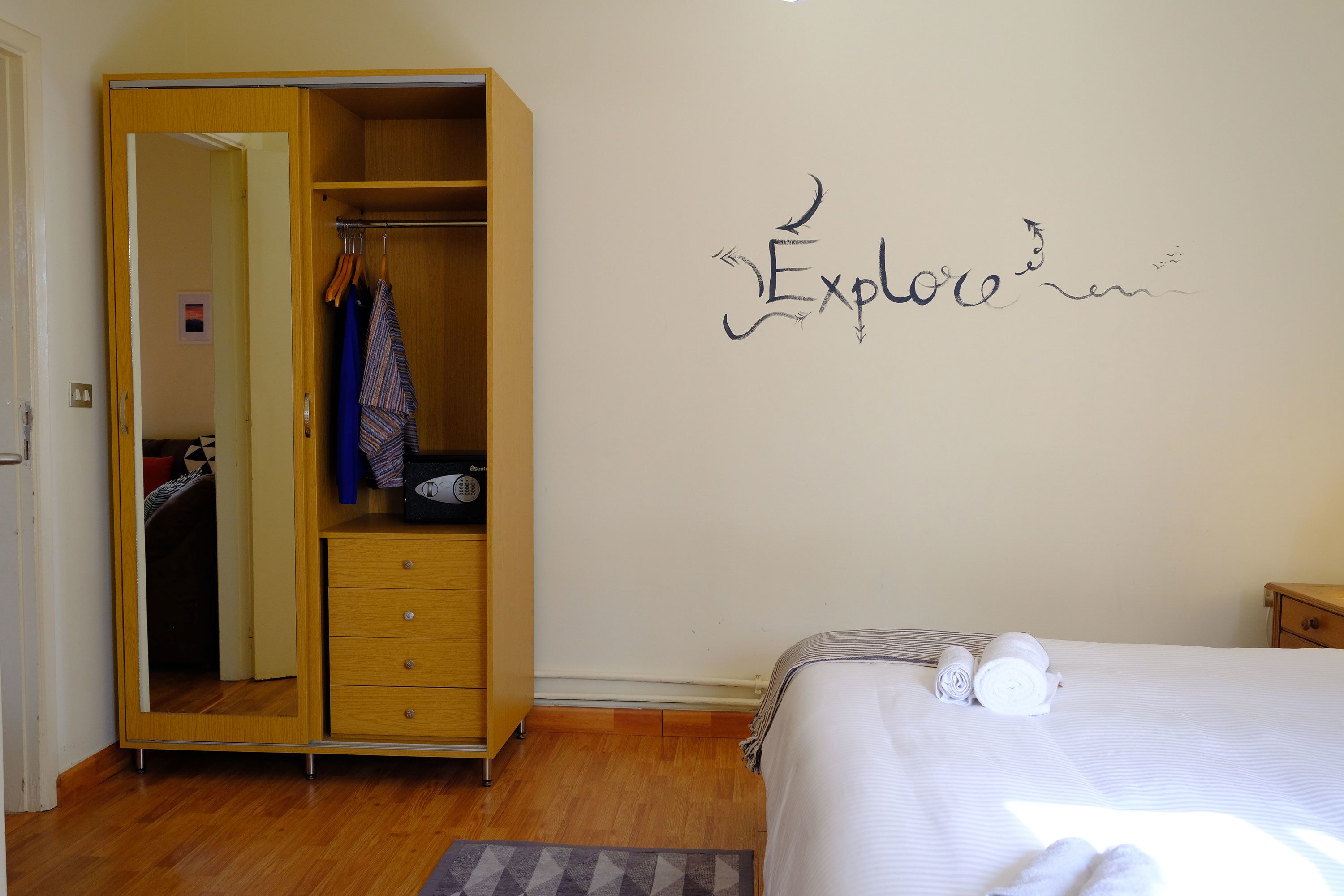 A bedroom with a wardrobe, a bed with towels, and a wall with the word "Explore" written on it.