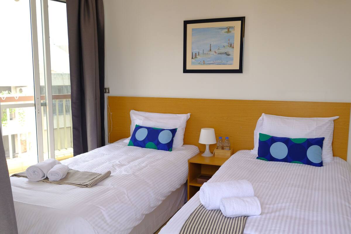 Ein Hotelzimmer mit zwei Betten, jeweils mit weißer Bettwäsche und blauen Kissen mit weißen Punkten. An der Wand über den Betten hängt ein gerahmtes Bild, neben jedem Bett ein kleiner Nachttisch mit Lampe und Flaschen, und ein Fenster mit Vorhängen.