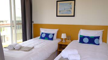 A hotel room with two beds, each with white bedding and blue pillows with white polka dots. There is a framed painting on the wall above the beds, a small wooden nightstand with a lamp and bottles, and a window with curtains.