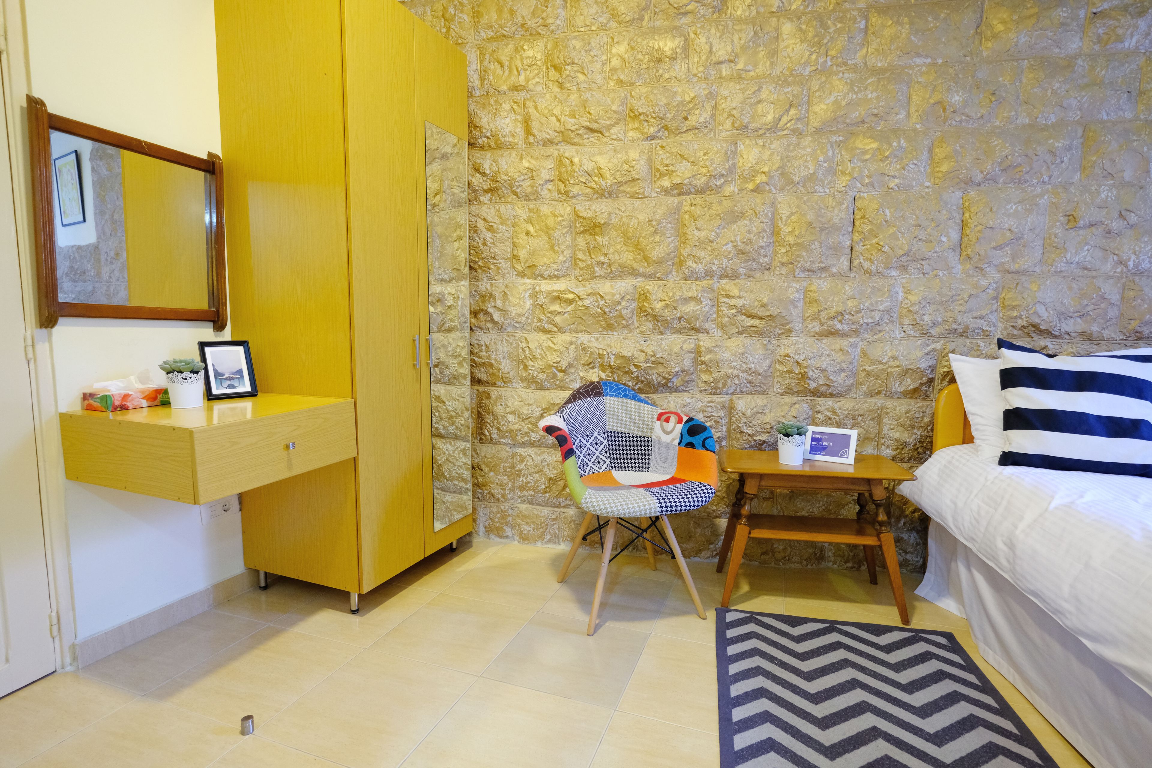 A room with a stone wall, a wooden wardrobe, a bed with striped bedding, a chair with a colorful cushion, and a small table with a plant.