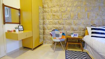 A room with a stone wall, a wooden wardrobe, a bed with striped bedding, a chair with a colorful cushion, and a small table with a plant.