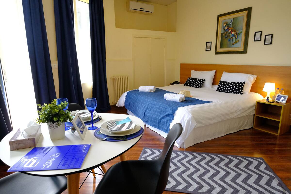 Ein Hotelzimmer mit Bett, Tisch mit Pflanze, Stuhl, Schreibtisch mit Computer und Fenster mit Vorhängen.