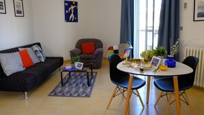 Una sala de estar con sofá, sillón, mesa de centro y mesa de comedor con sillas. Hay una ventana con cortinas, un radiador y un cuadro en la pared.