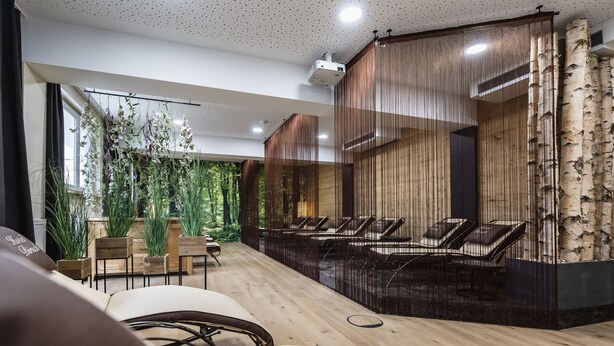 Un espacio interior moderno con suelo de madera, una zona de estar con sillas, un ventanal con plantas y una pared con listones de madera verticales.