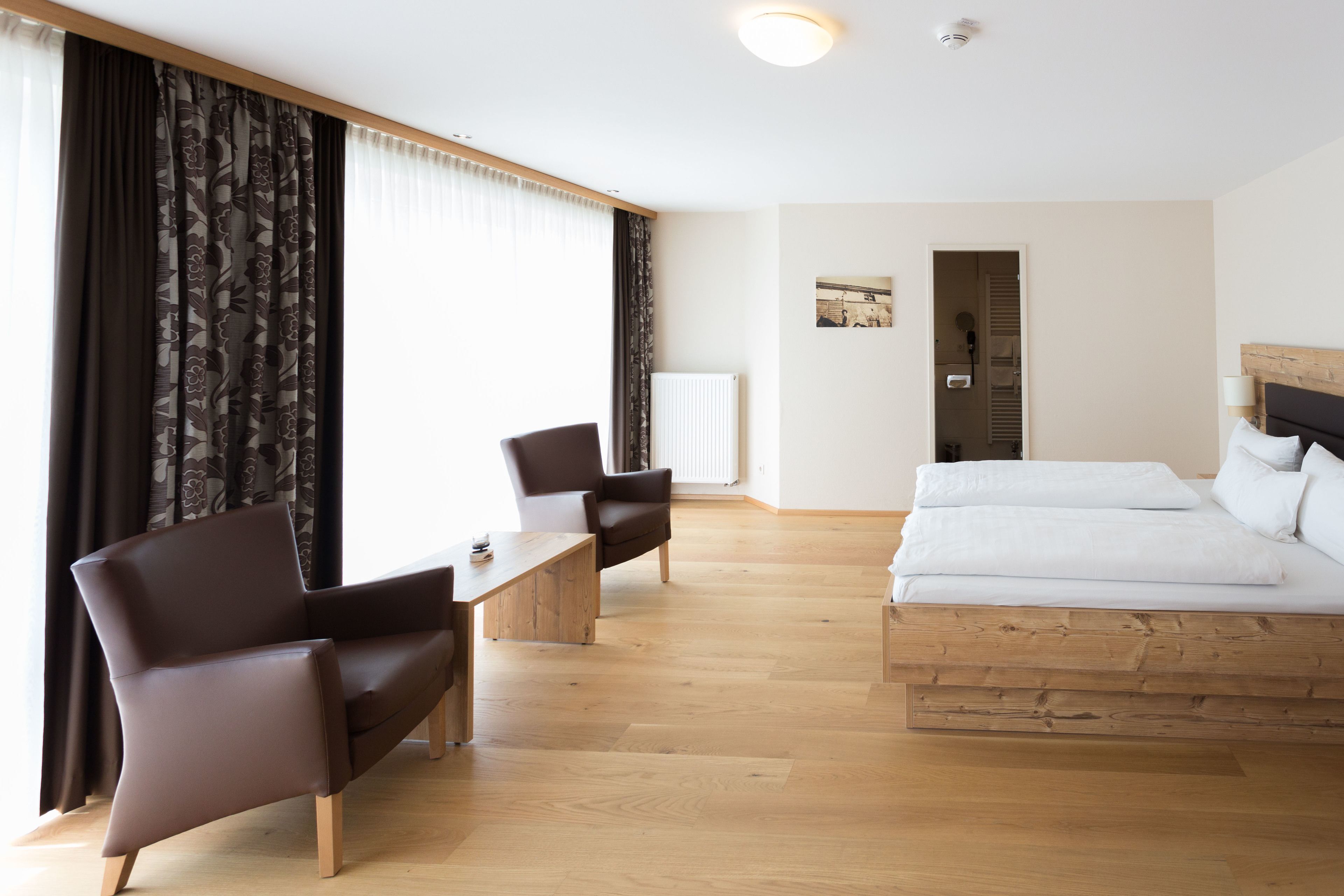 A modern hotel room with a wooden bed, a desk, a chair, and a large window with curtains.