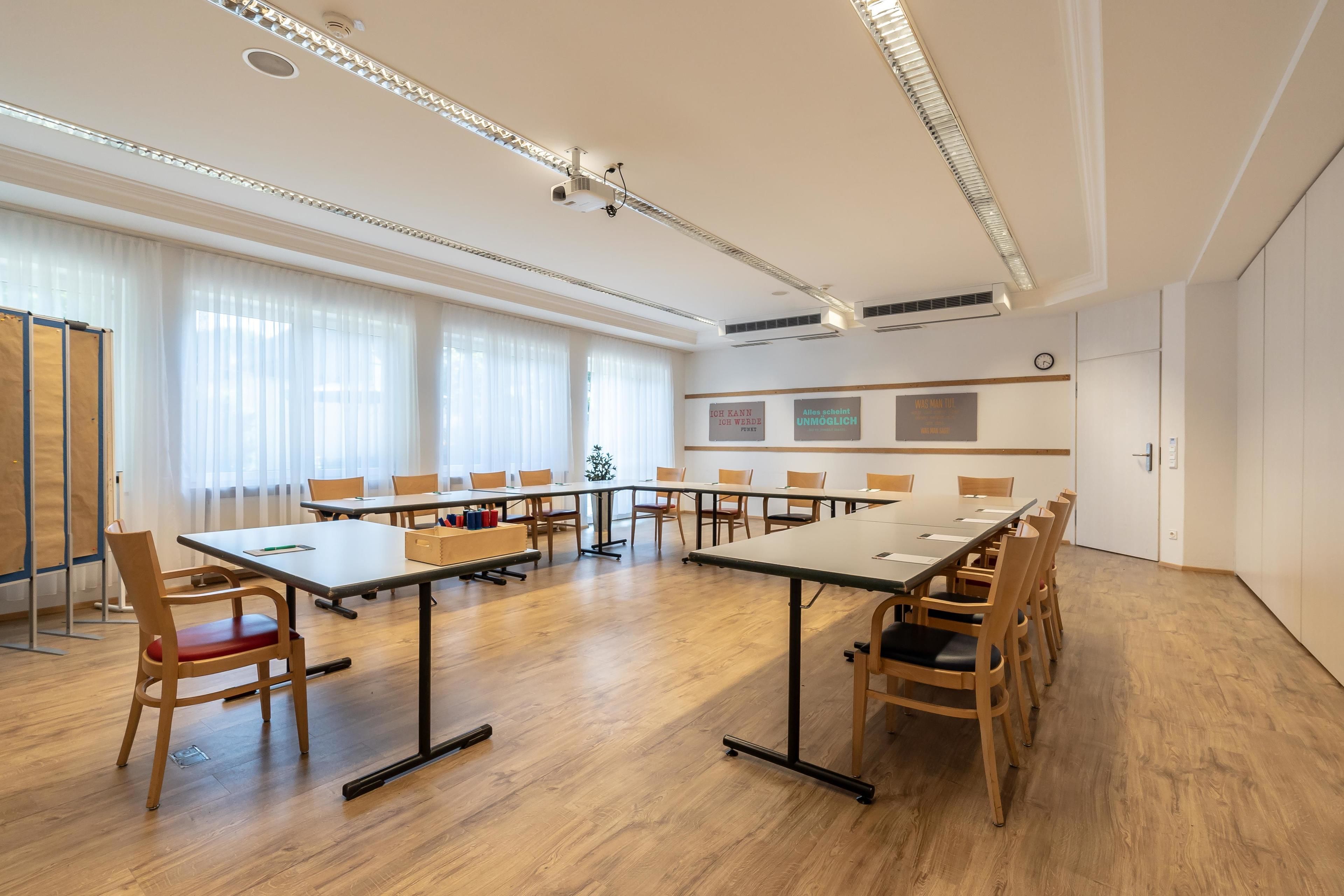 Una sala conferenze con lunghe tavolate e sedie, ampie finestre con tende leggere e un soffitto bianco con illuminazione a incasso.