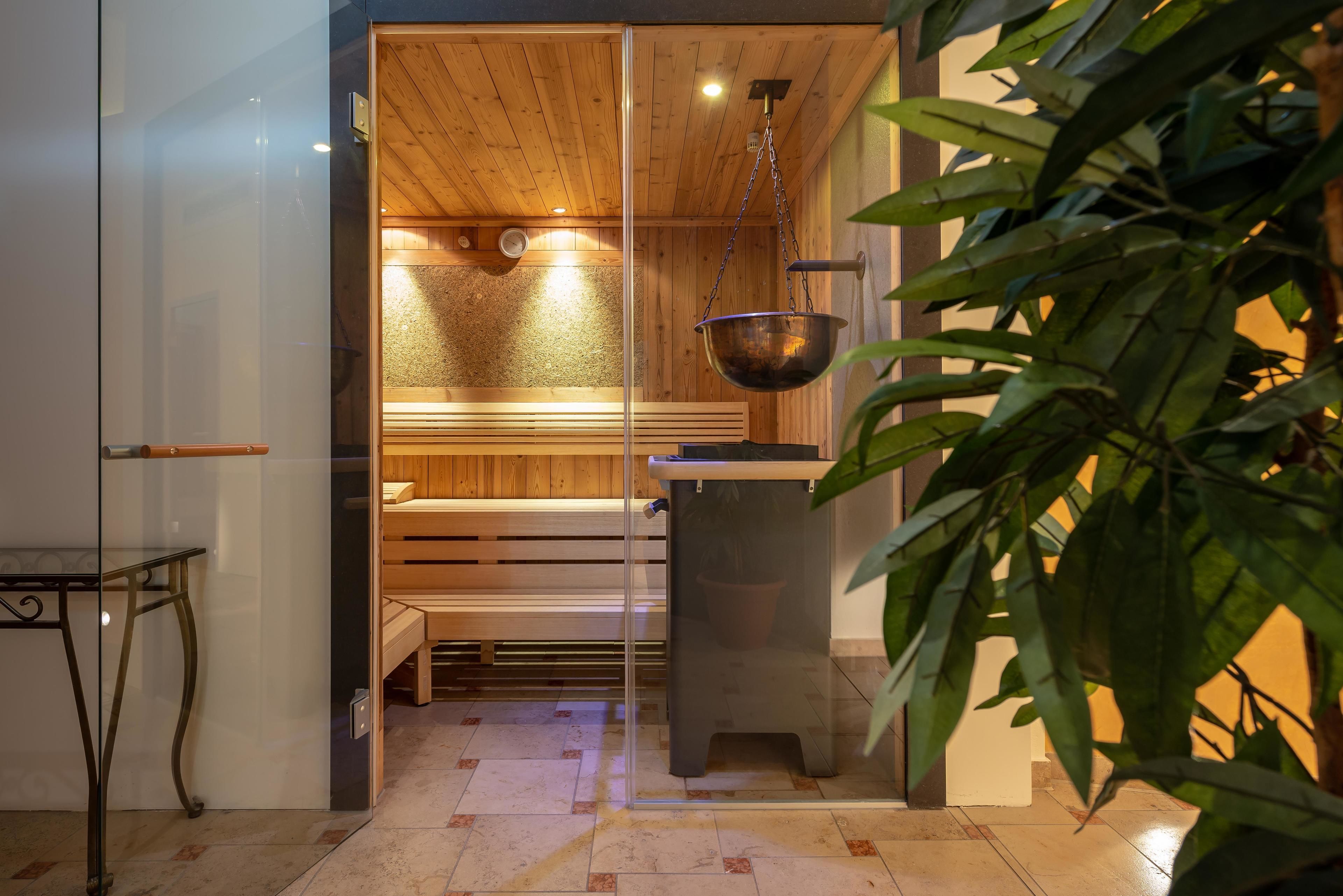 A sauna room with wooden benches, a hanging metal basin, and a glass partition.