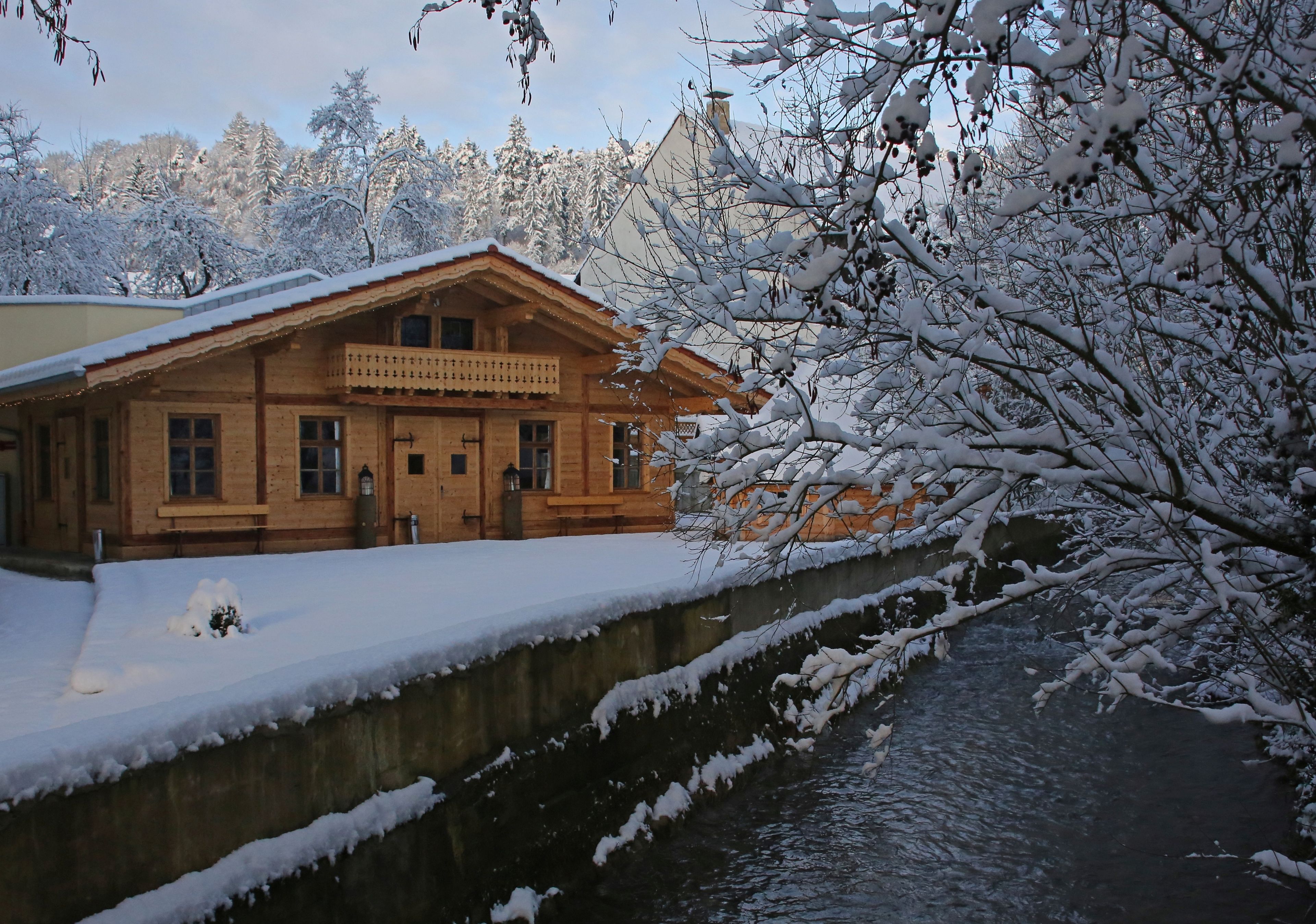 Ein Holzgebäude mit Balkon, ein schneebedeckter Fluss und Bäume im Hintergrund.