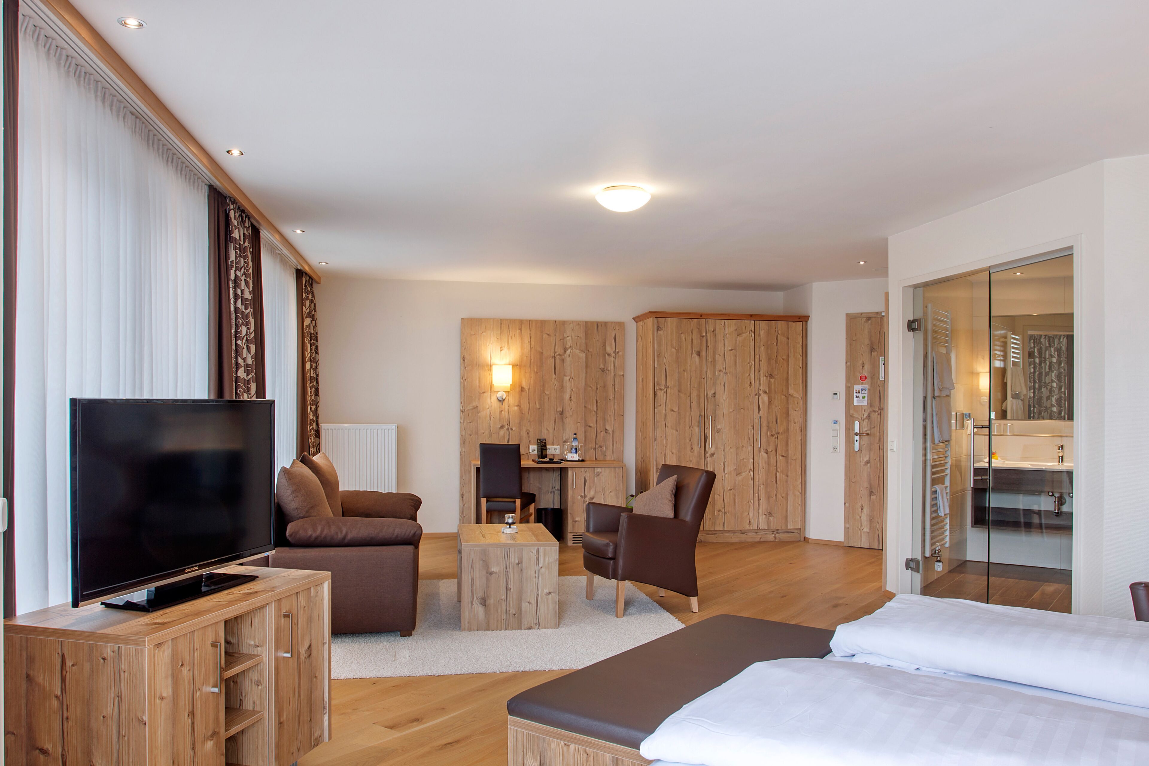 A modern hotel room with a flat-screen TV, a sofa, a chair, a small wooden table, and a bed.