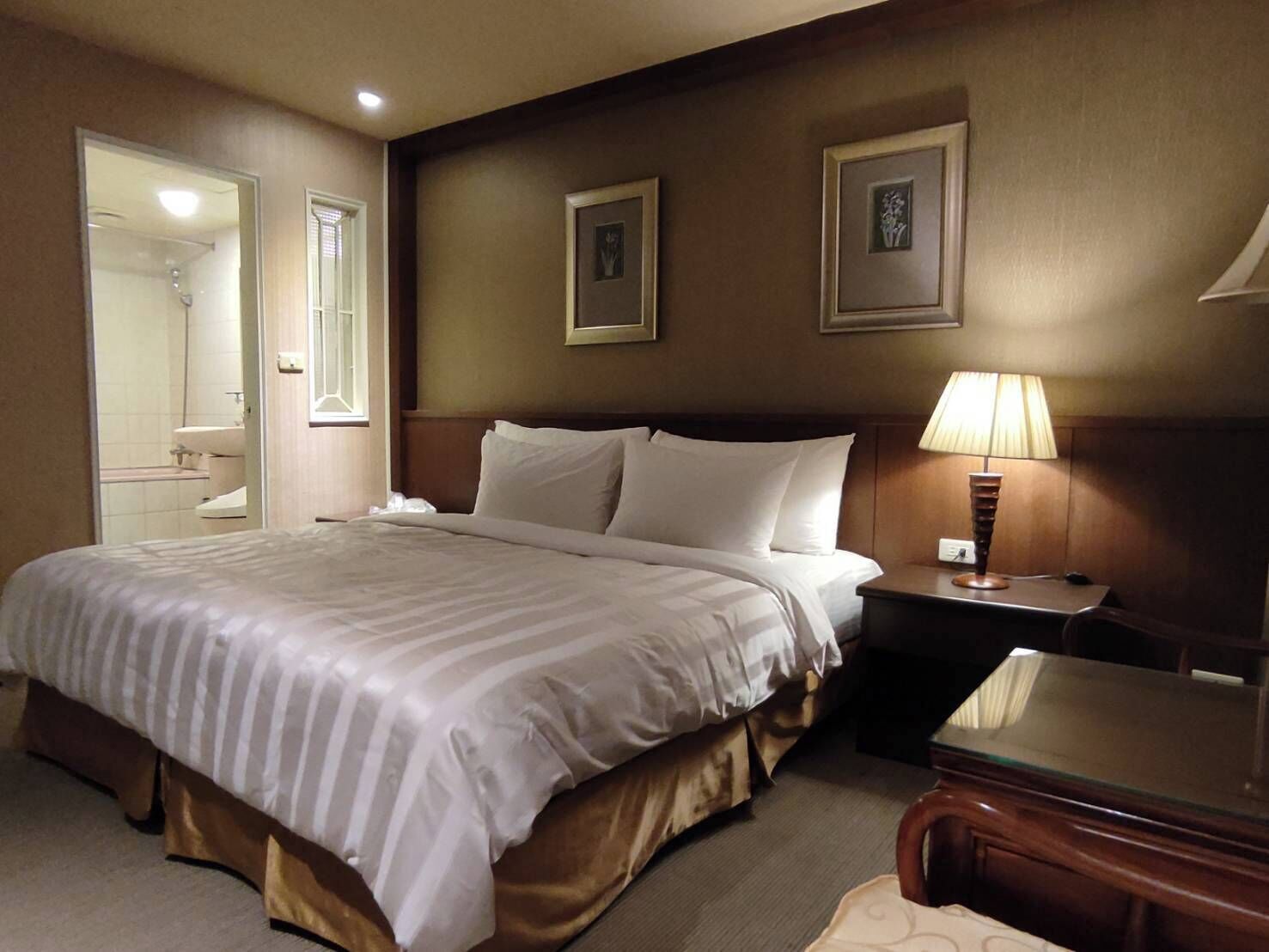 Une chambre d’hôtel avec un grand lit, une table de chevet avec une lampe, une commode en bois et des tableaux encadrés au mur.