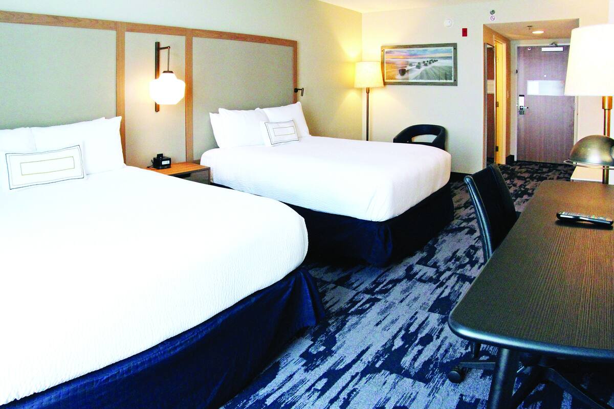 A hotel room with two beds, a desk, and a chair. There is a lamp on the desk and a painting on the wall.