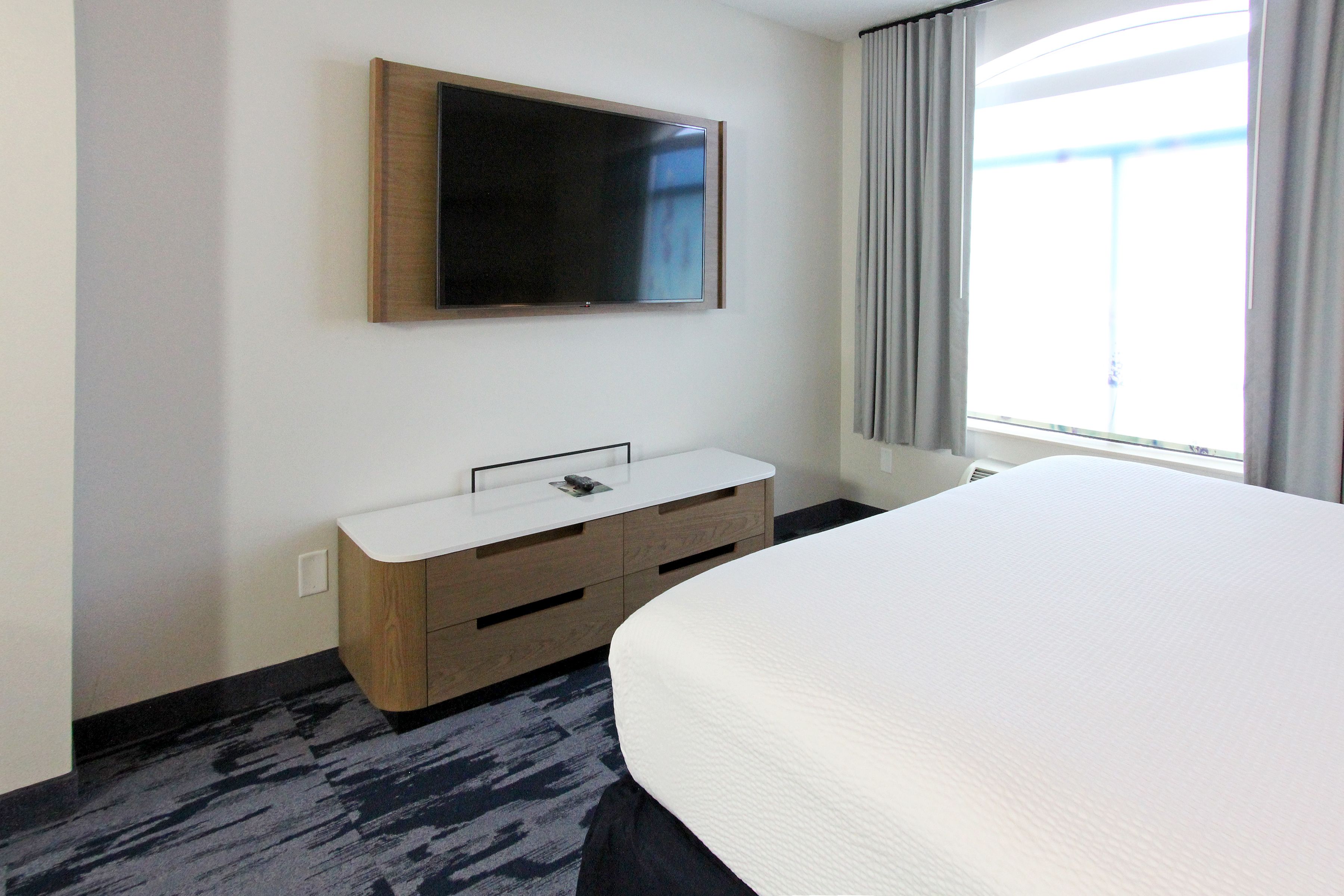 A hotel room with a large bed, a flat-screen TV mounted on the wall, a wooden dresser, and a window with curtains.