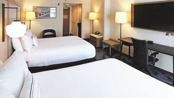 A hotel room with two beds, a desk, a chair, a TV, and a lamp.