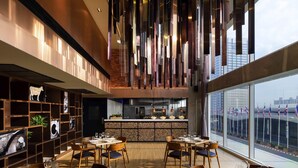 A modern restaurant with high ceilings, hanging light fixtures, and a view of the cityscape through large windows. - Novotel Bangkok IMPACT (Pak Kret)