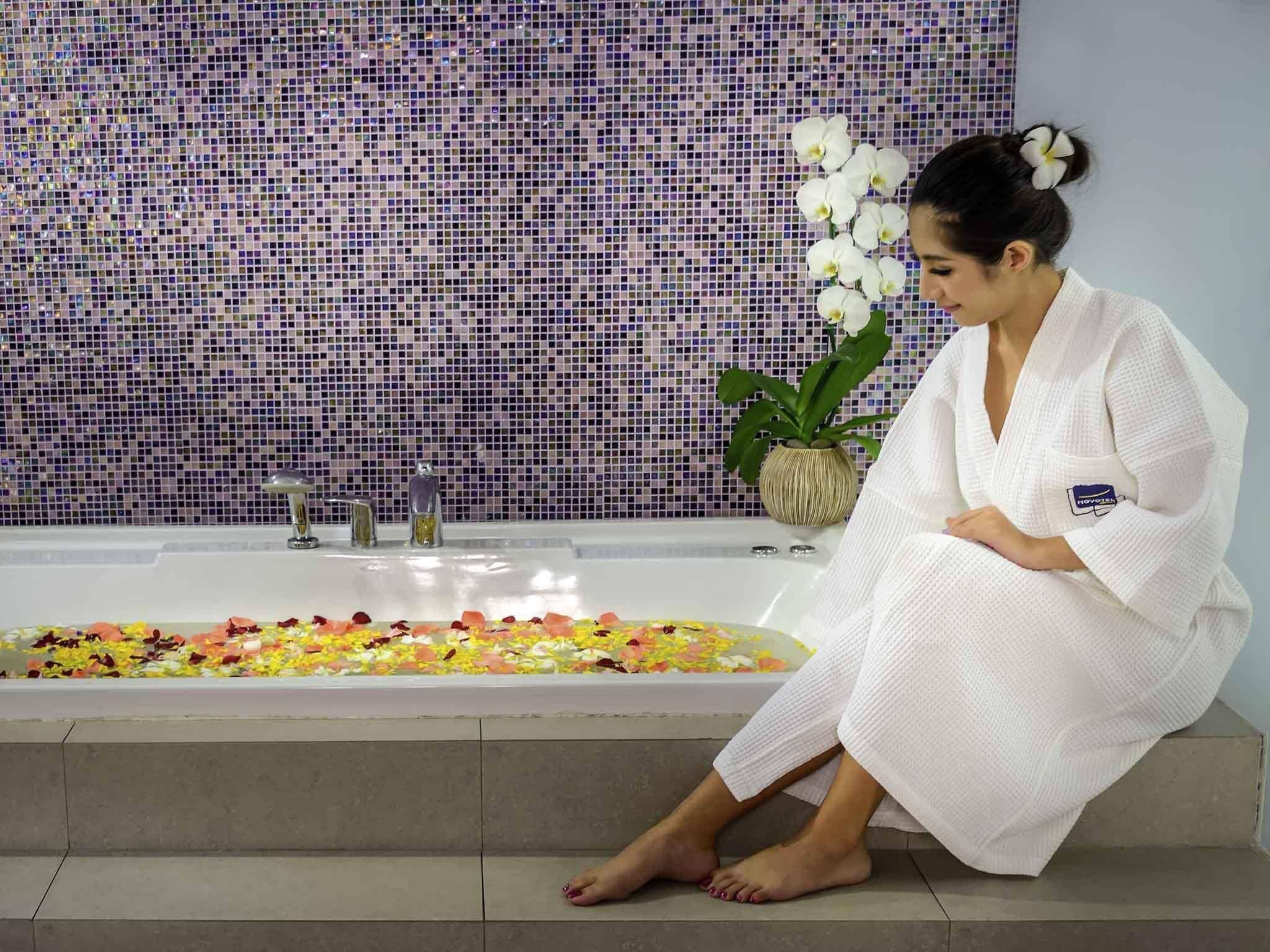 A person sitting by a bathtub filled with flower petals, with a mosaic tile wall in the background.