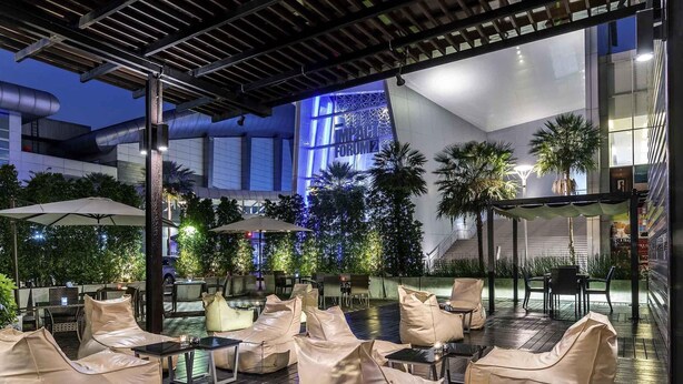 An outdoor seating area with cushioned chairs and tables, surrounded by greenery and a modern building in the background. - Novotel Bangkok IMPACT (Pak Kret)