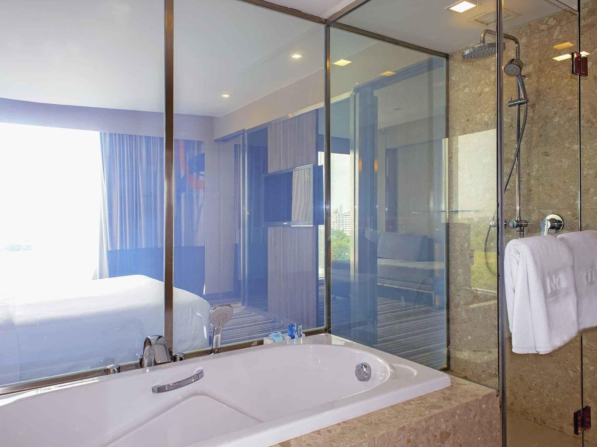 Une salle de bain d’hôtel moderne, dotée d’une grande baignoire, d’une douche en verre et d’un lit visible dans la pièce adjacente.
