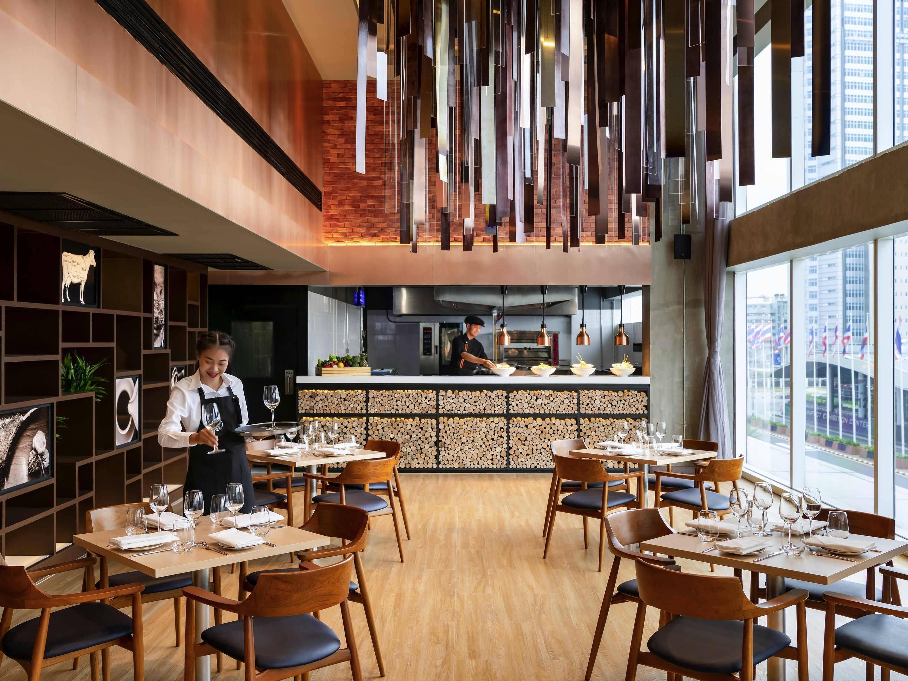 Ein Restaurant mit Holztischen, einem Koch der kocht und Blick auf die Stadt durch große Fenster.