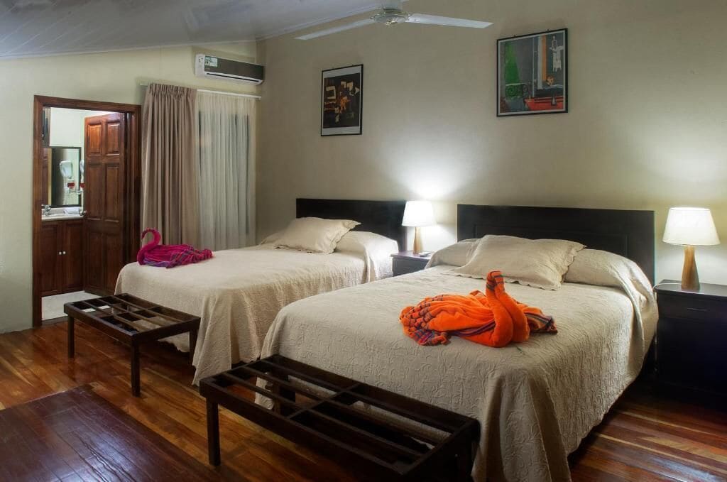 A hotel room with two beds, a wooden bench, a ceiling fan, and a window with curtains.