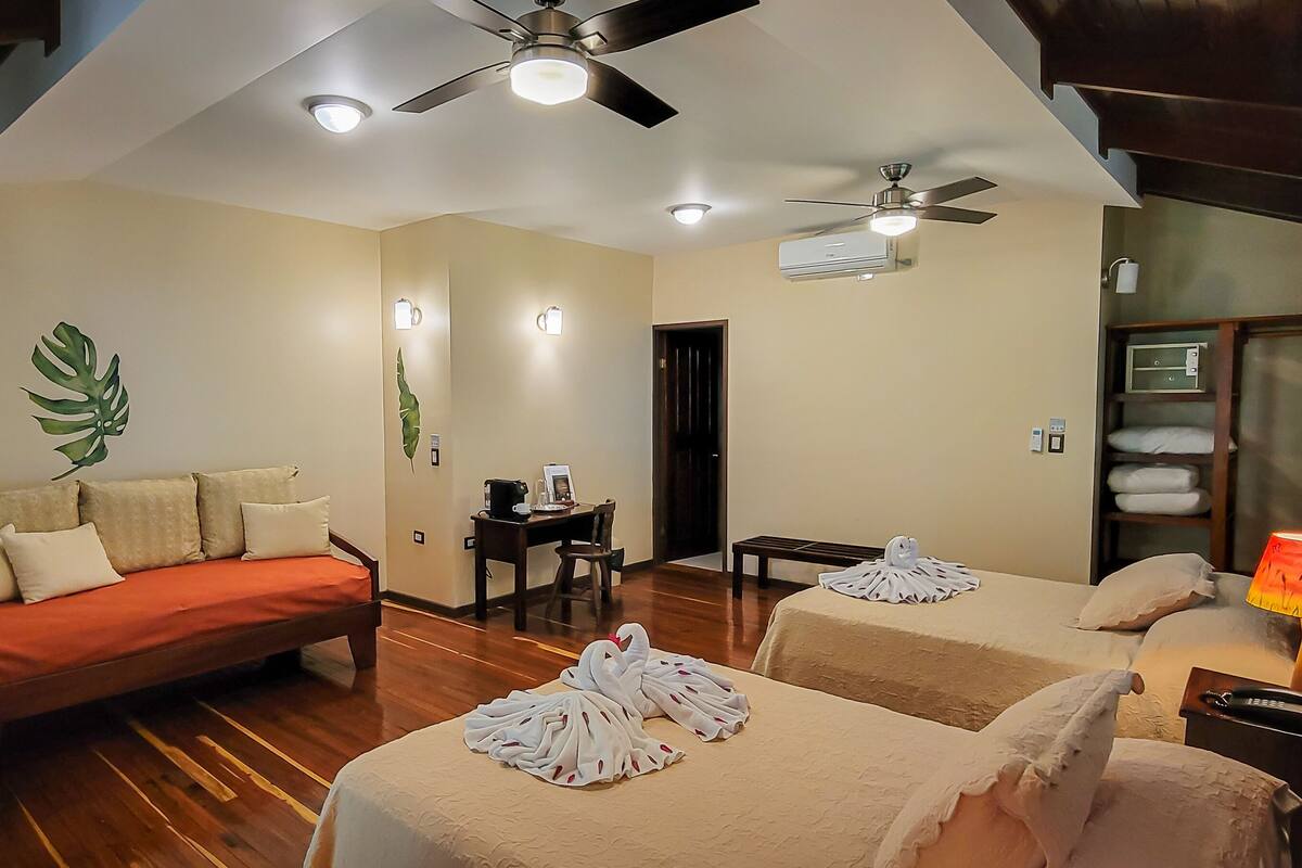 A hotel room with a sofa, two beds, a desk, a ceiling fan, and a nightstand with a lamp.