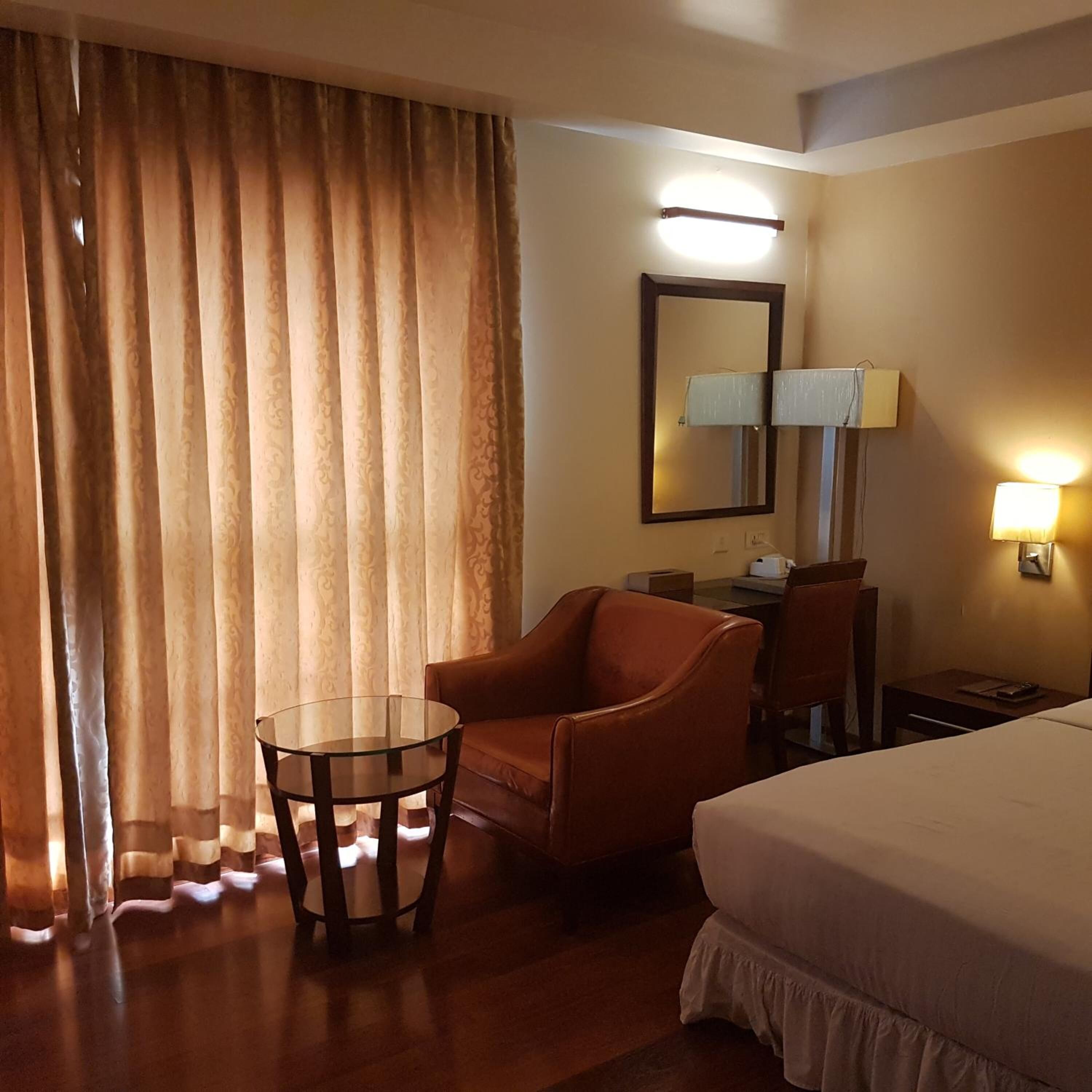 A hotel room with a bed, a chair, a small table, a desk, a lamp, and a mirror.