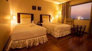 A hotel room with two beds, a small table, and a view of the city at dusk.