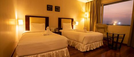 Habitación de hotel con dos camas, una mesita y vista a la ciudad al anochecer.